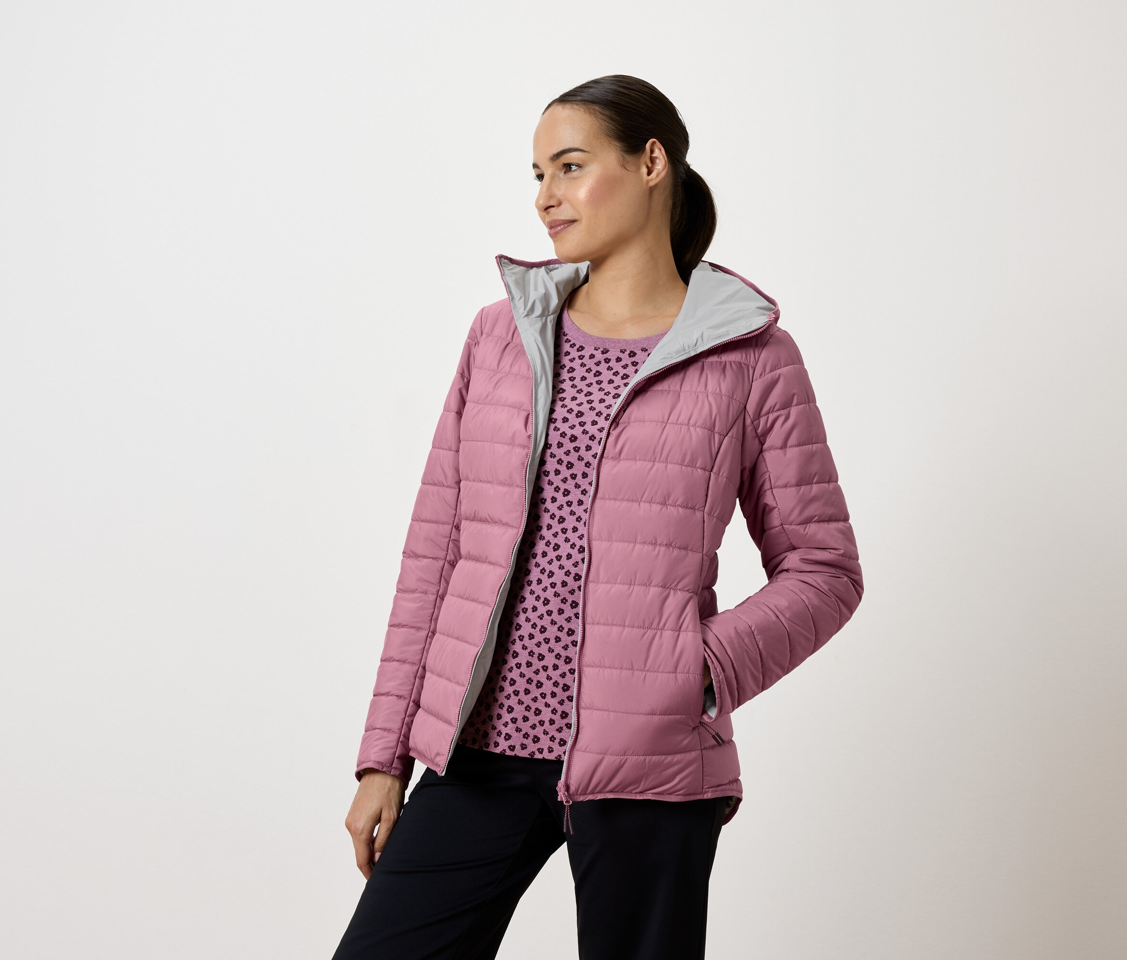 Žena v ružovej prešívanej bunde s kapucňou a tmavomodrých nohaviciach, pozerá sa doľava.