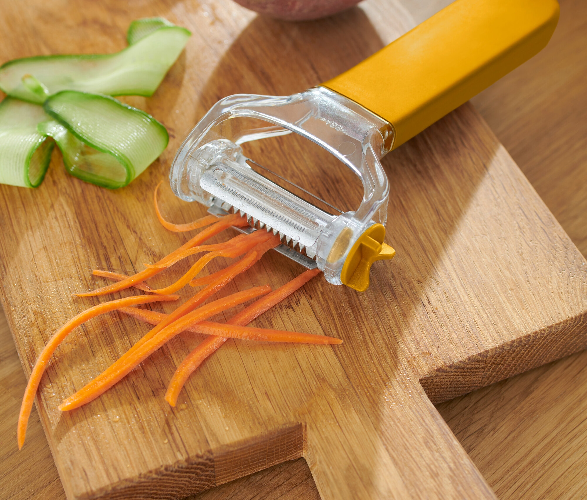 Žltá škrabka na zeleninu s ošúpanou mrkvou, uhorkou a zemiakom na drevenej doske.