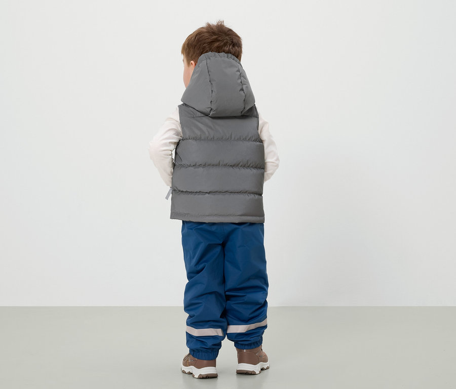 Chlapec stojí chrbtom, má na sebe sivú vestu s kapucňou, modré nohavice a hnedé topánky.