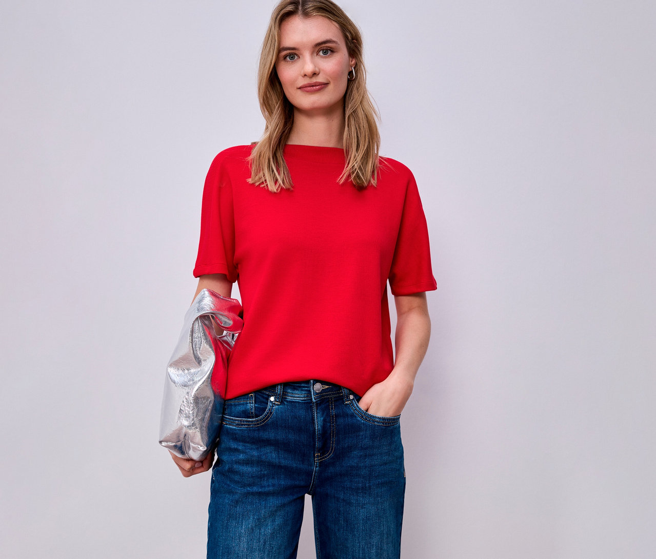 Žena v červenom tričku s krátkym rukávom Street One a džínsoch, cez rameno má prehodenú striebornú bundu.