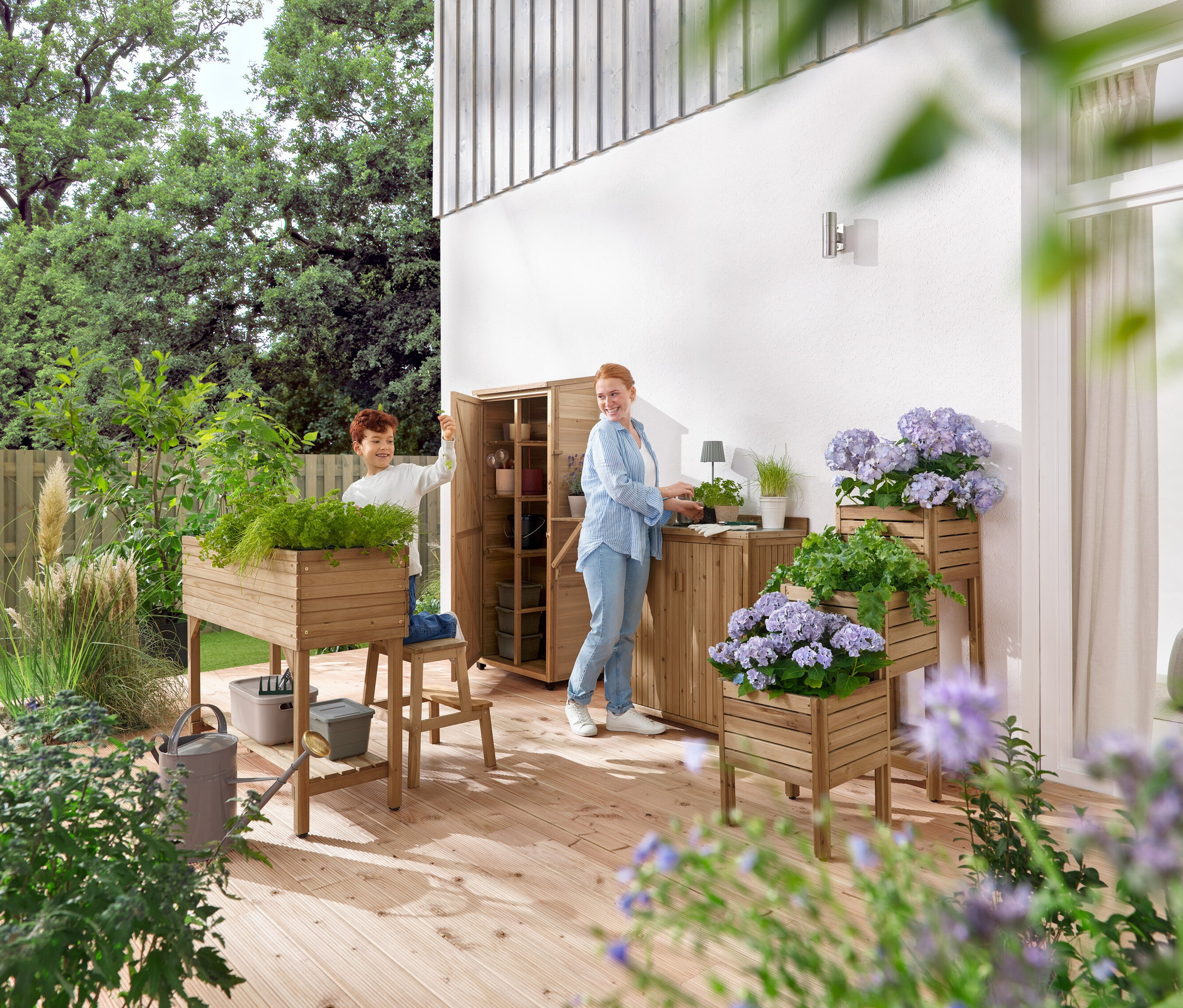 Chlapec a žena pracujú na drevenej terase so záhradným nábytkom a rastlinami.