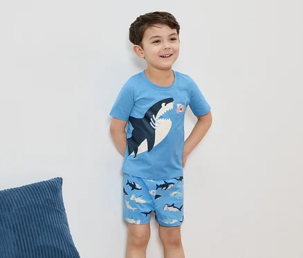 Usmievavý chlapec má na sebe svetlomodré pyžamo s motívom žraloka, stojí pred bielou stenou.