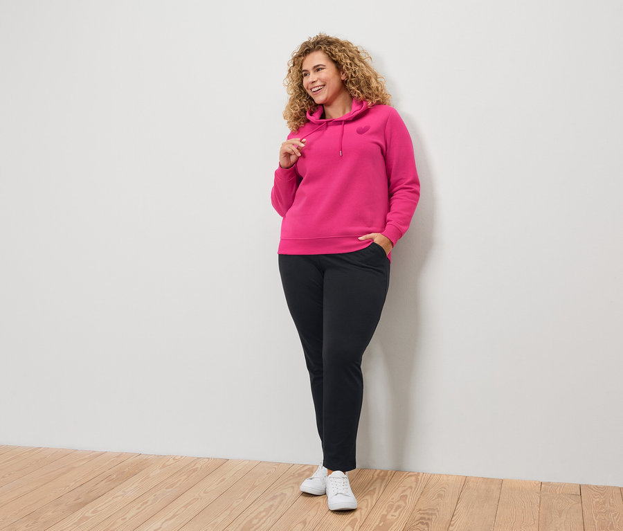 Žena s kučeravými vlasmi v ružovej mikine s kapucňou a čiernych nohaviciach.