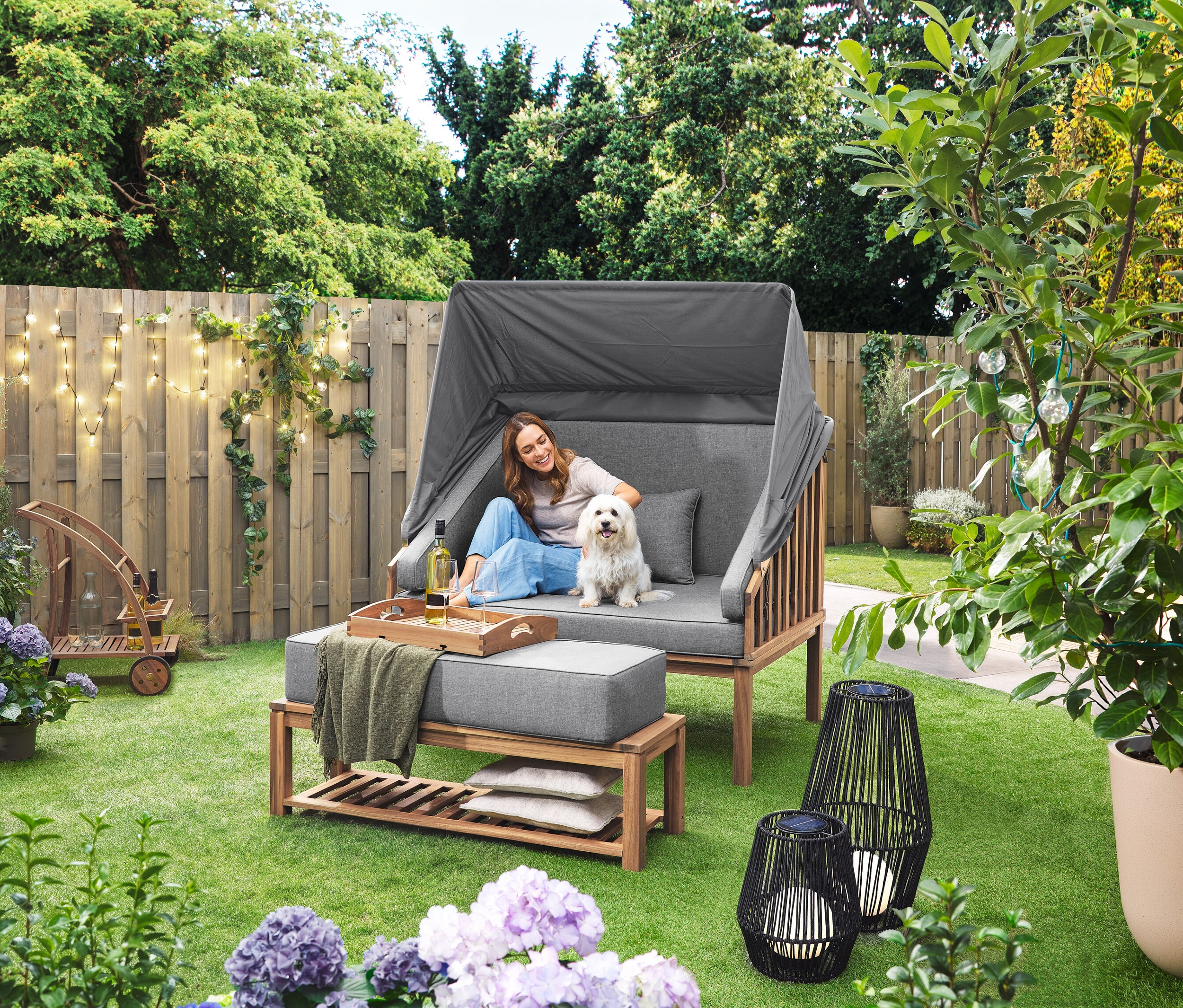 Žena sedí so psom v plážovom koši Premium Lounge-Strandkorb mit Sonnenschutz-Dach. Na trávniku stojí solárny lampáš s textilnou šnúrkou, veľký a solárny lampáš s textilnou šnúrkou, malý. V pozadí je barový vozík s odnímateľnou táckou. Na plote visí LED-Outdoor-Lichterkette mit Netzadapter.