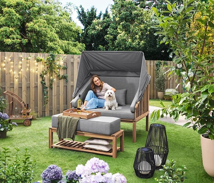 Žena sedí so psom v plážovom koši Premium Lounge-Strandkorb mit Sonnenschutz-Dach. Na trávniku stojí solárny lampáš s textilnou šnúrkou, veľký a solárny lampáš s textilnou šnúrkou, malý. V pozadí je barový vozík s odnímateľnou táckou. Na plote visí LED-Outdoor-Lichterkette mit Netzadapter.
