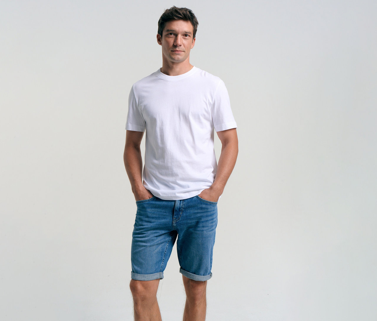 Muž v bielom tričku a džínsových šortkách Tom Tailor TTJOSH REGULAR SLIM, mid stone bright blue denim, s rukami vo vreckách.
