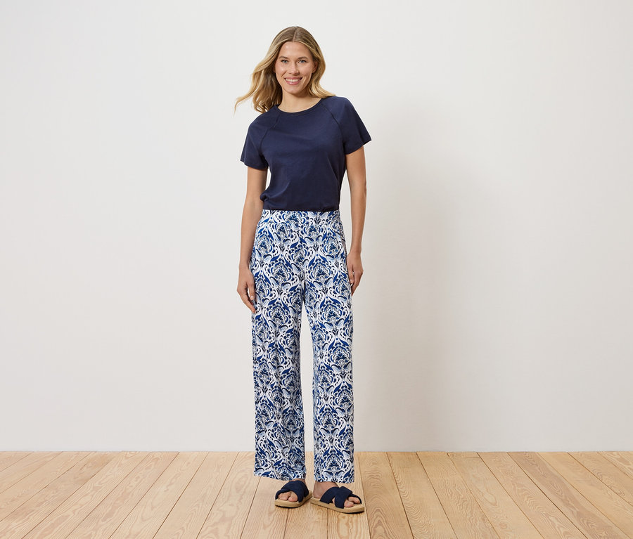 Žena stojí pred bielou stenou a má na sebe tmavomodré tričko, modré a biele vzorované nohavice a modré sandále.