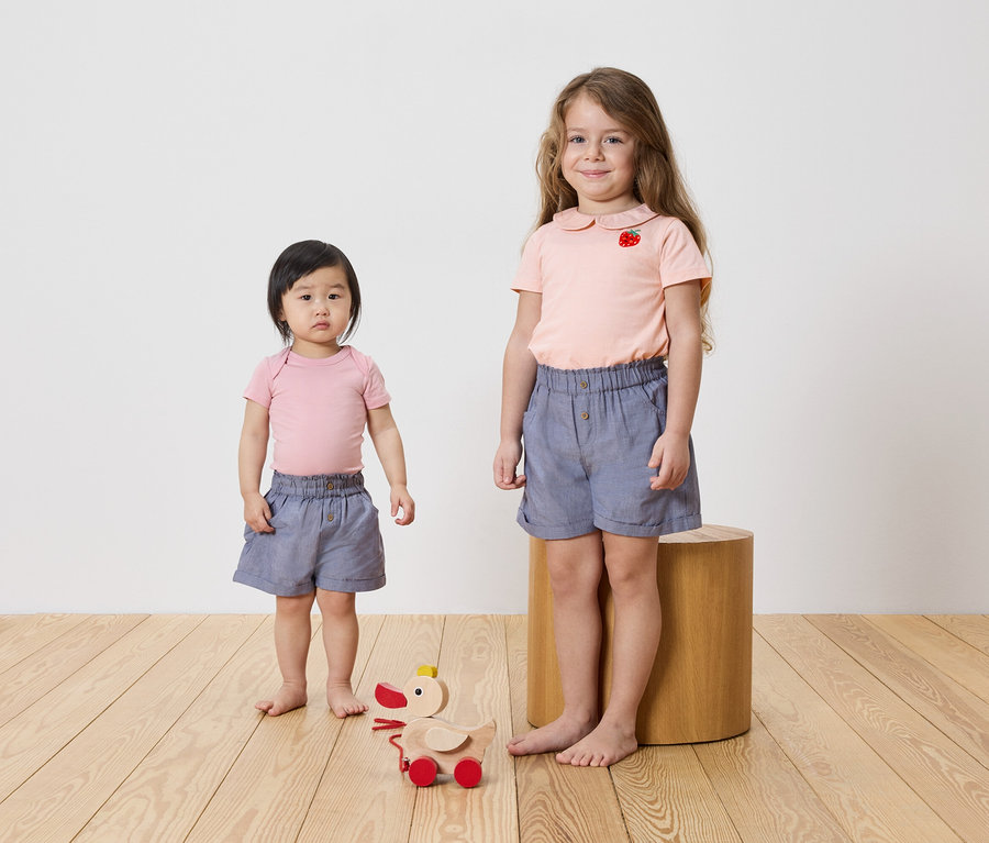 Dve dievčatá stoja na drevenej podlahe, obe majú na sebe ružové topy a modré šortky. Na podlahe je drevená hračka.