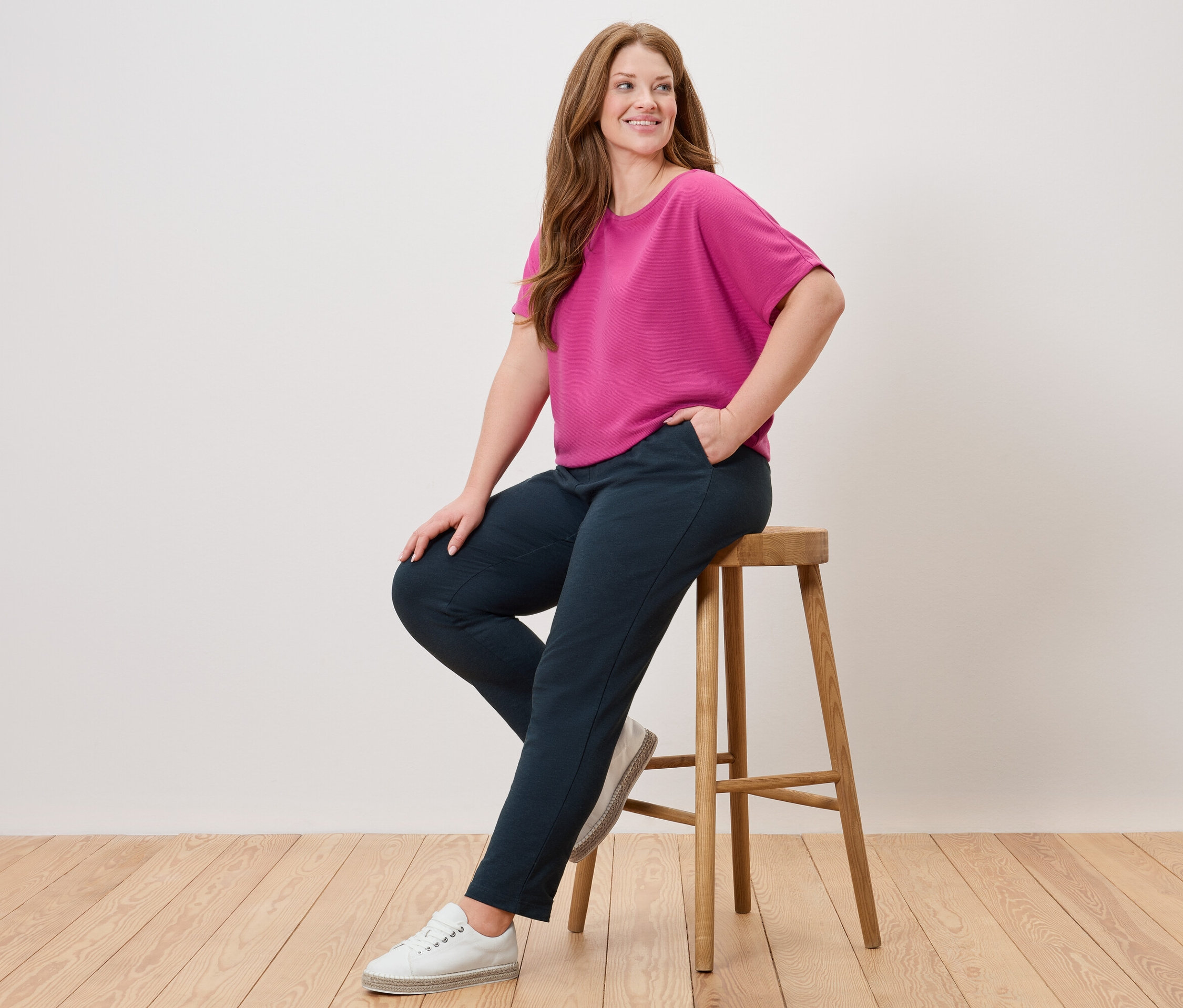 Žena sedí na stoličke, má na sebe ružovú blúzku, modré nohavice a biele topánky.