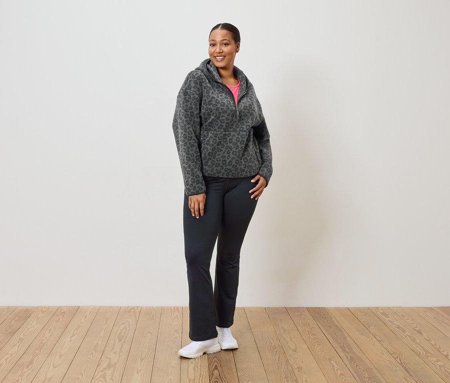 Žena stojí a má na sebe sivo-čiernu leopardiu flísovú mikinu s kapucňou, čierne nohavice a biele tenisky.