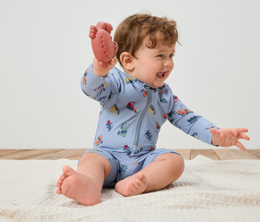 Smejúce sa bábätko s kučeravými vlasmi sedí na bielej deke a v ruke drží červené hryzátko. Bábätko má na sebe svetlomodrý overal s farebnými vozidlami.