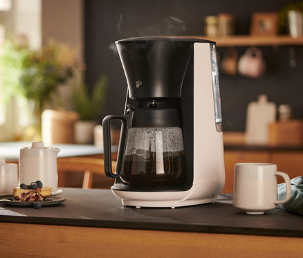 Biely kávovar na prekvapkávanú kávu »Let's Brew« stojí na stole vedľa šálky, kanvičky na mlieko, cukorničky a taniera s koláčom.