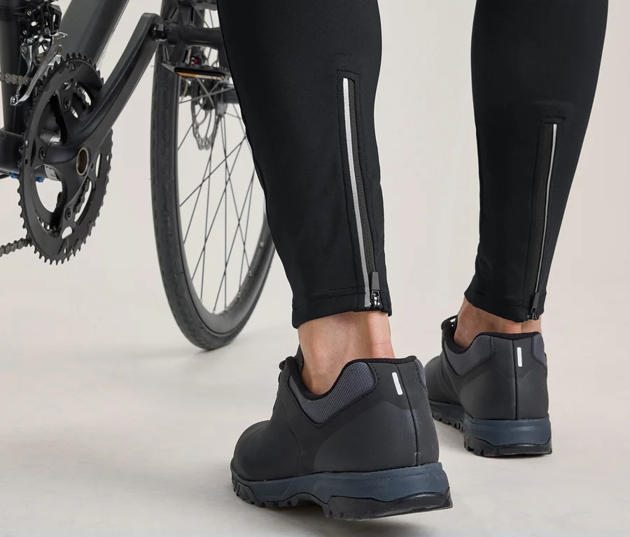 Spodné časti nôh a chodidlá osoby stojacej vedľa bicykla. Osoba má na sebe čierne nohavice s reflexnými zipsami na členkoch a čierne cyklistické topánky.