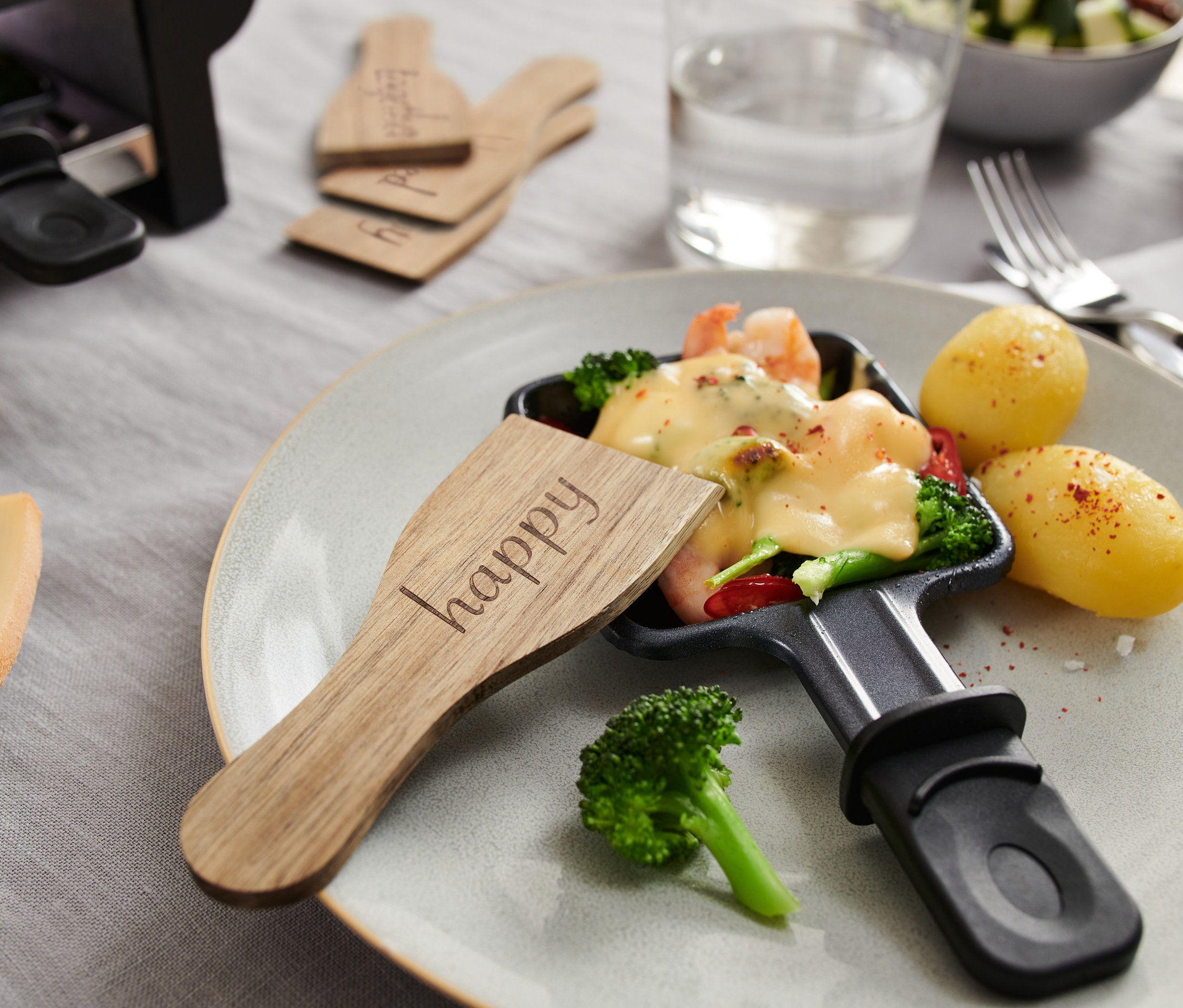 Detailný záber na raclette panvicu s roztaveným syrom, zeleninou a zemiakmi, s drevenou lopatkou na raclette (4 ks) s nápisom "happy".