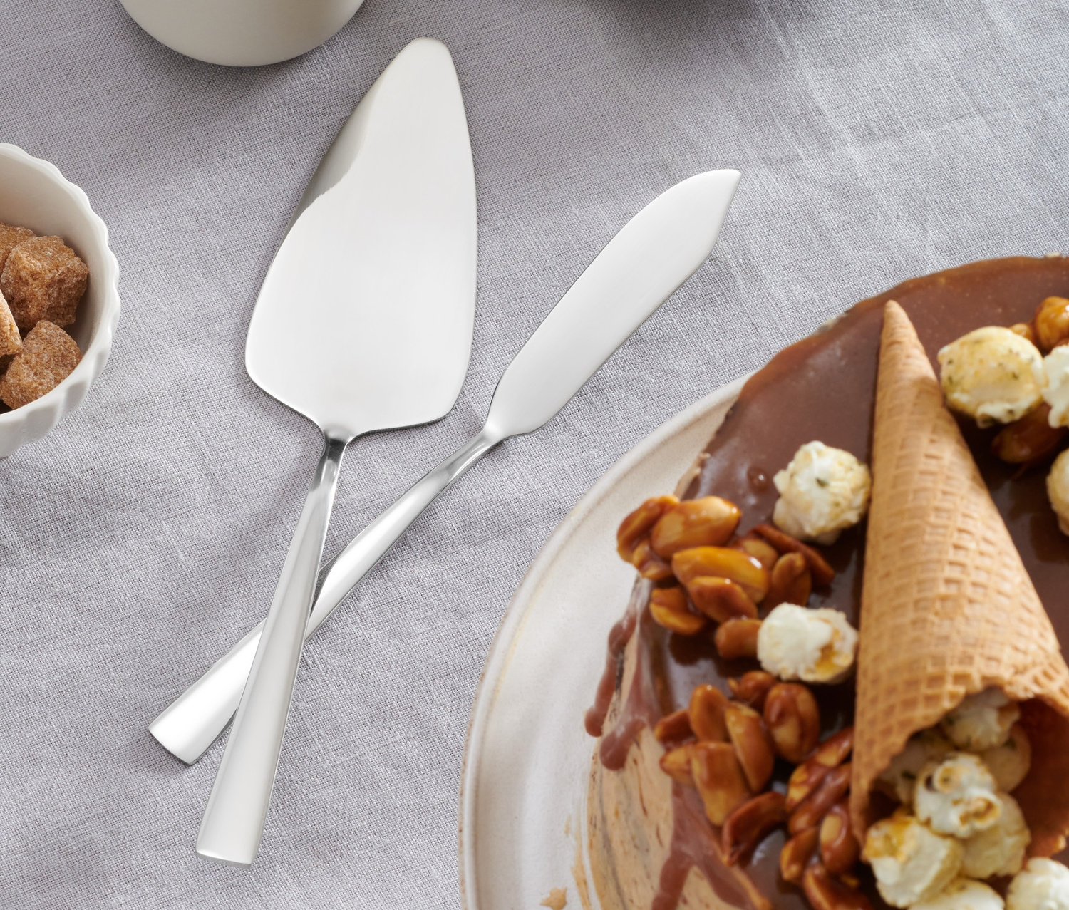 Na stole leží torta s polevou, orechmi a popcornom vedľa 2-dielnej súpravy lopatky a noža na tortu.