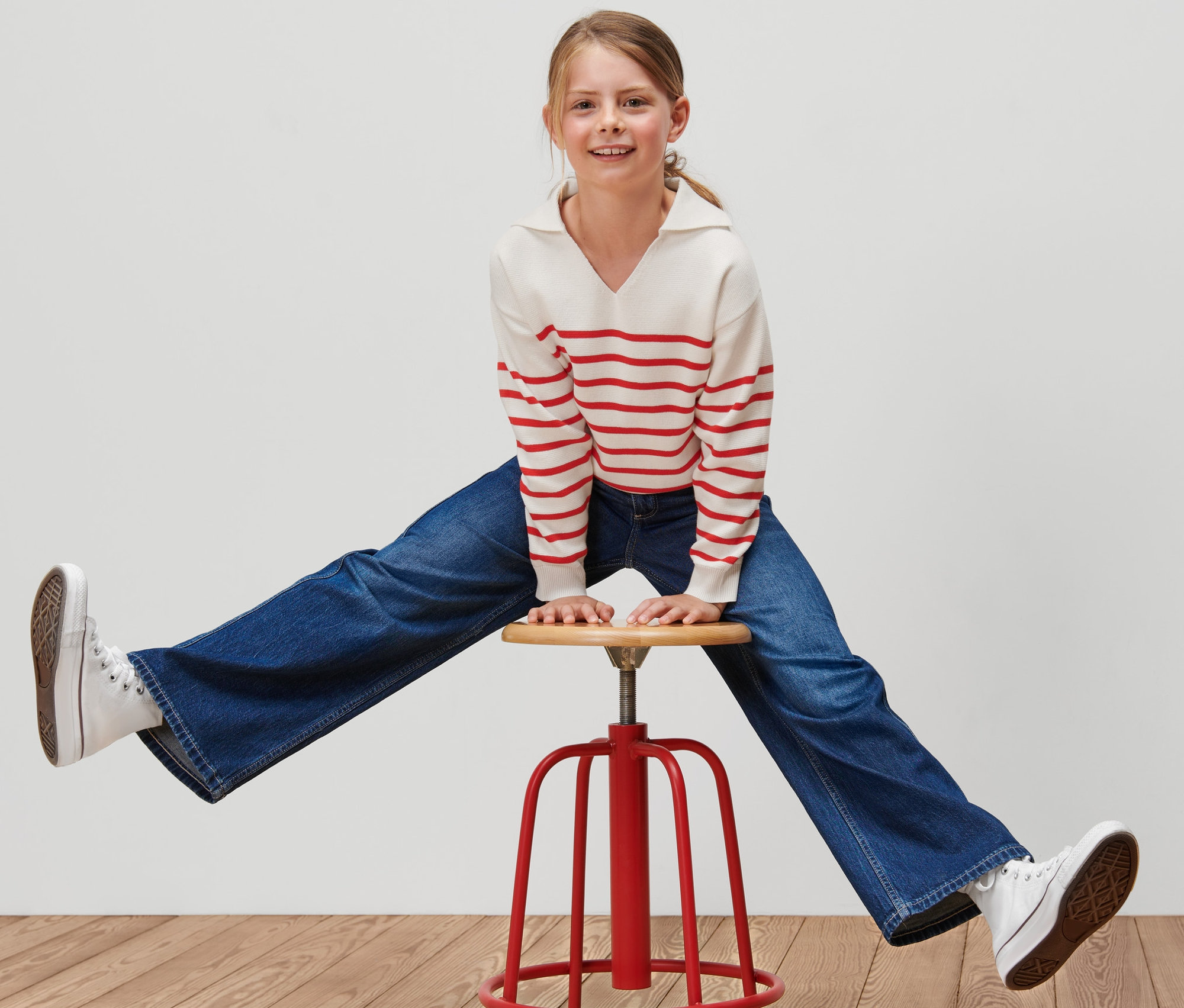 Dievča sedí rozkročené na červenej stoličke a má na sebe detský pletený pulóver s červenými pruhmi, modré džínsy a biele tenisky.