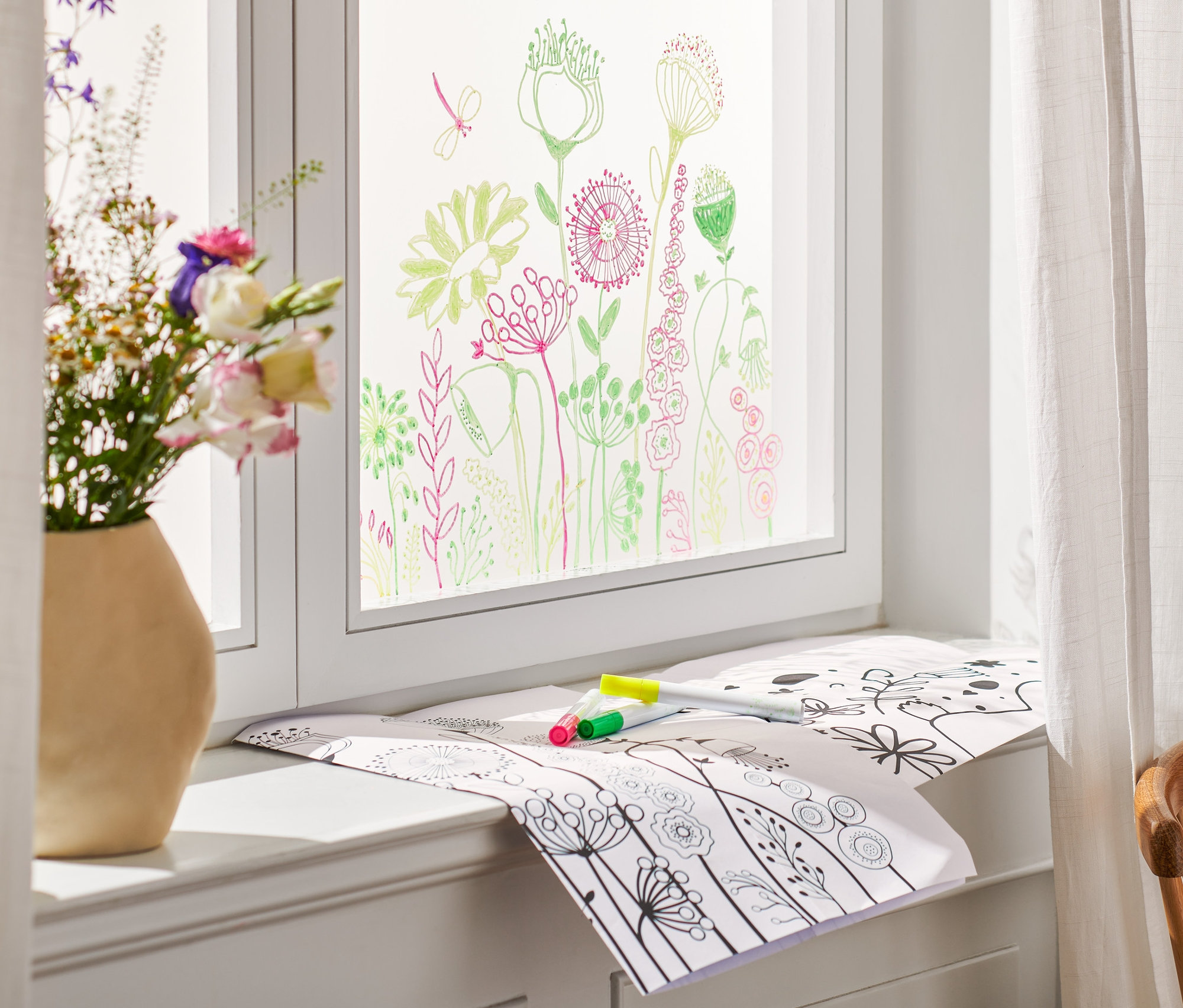 Kvety vo váze stoja na okennom parapete, okno je pomaľované a na parapete ležia Veľkonočná maľovanka na dekoráciu okien a fixky.