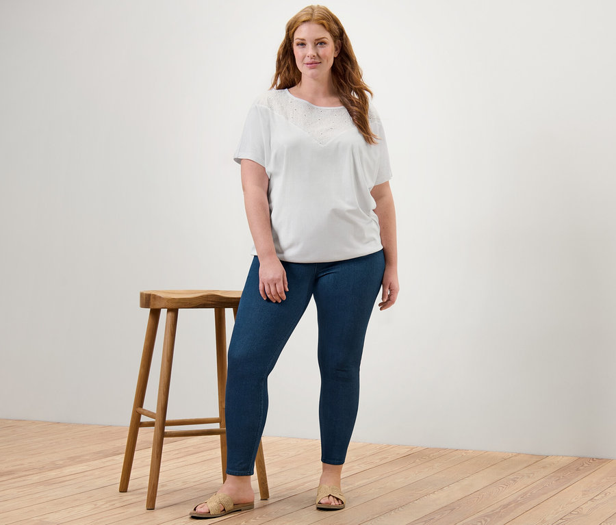 Žena stojí a má na sebe biele blúzkové tričko s výšivkou a džegínsy.