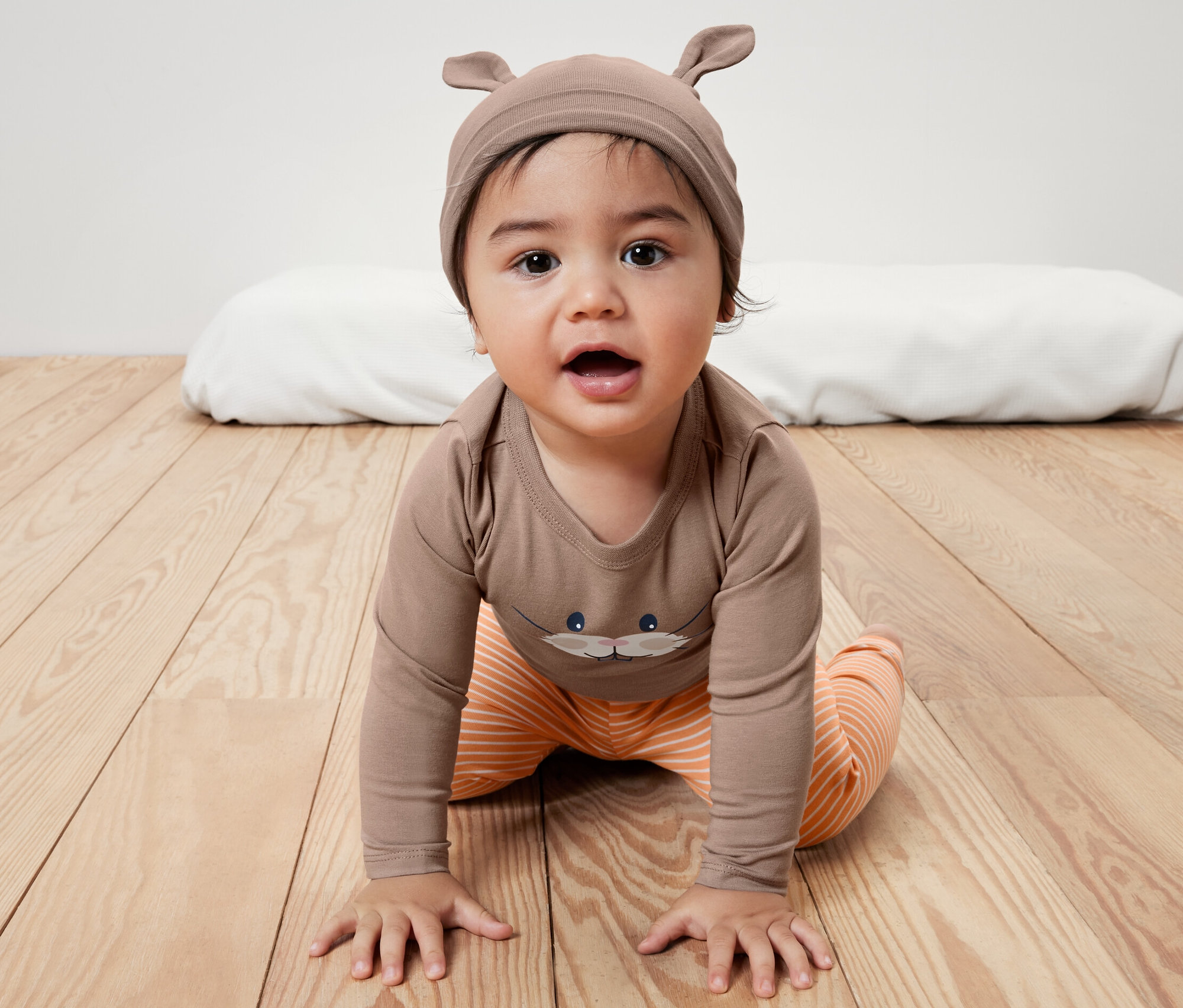 Súprava pre bábätko: Dieťa lezie po drevenej podlahe v hnedom body s potlačou a oranžových pruhovaných nohaviciach. Hnedá čiapka s ušami.