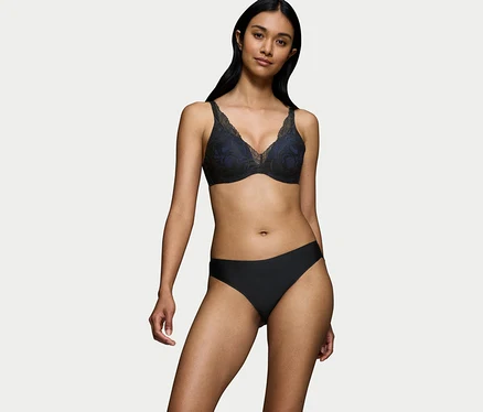 Žena s tmavými vlasmi v čiernej podprsenke Triumph Body Makeup Illusion Lace WP s modrou podšívkou a čiernych nohavičkách, stojí pred bielym pozadím.