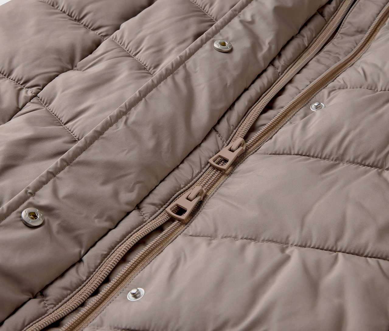 Detail béžovej prešívanej bundy so zipsom a patentkami.