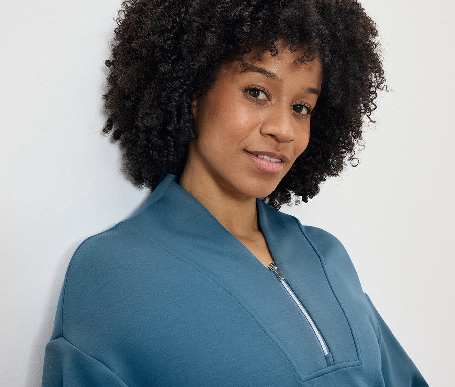 Portrét usmievajúcej sa ženy s afro účesom v modrozelenej mikine so zipsom.