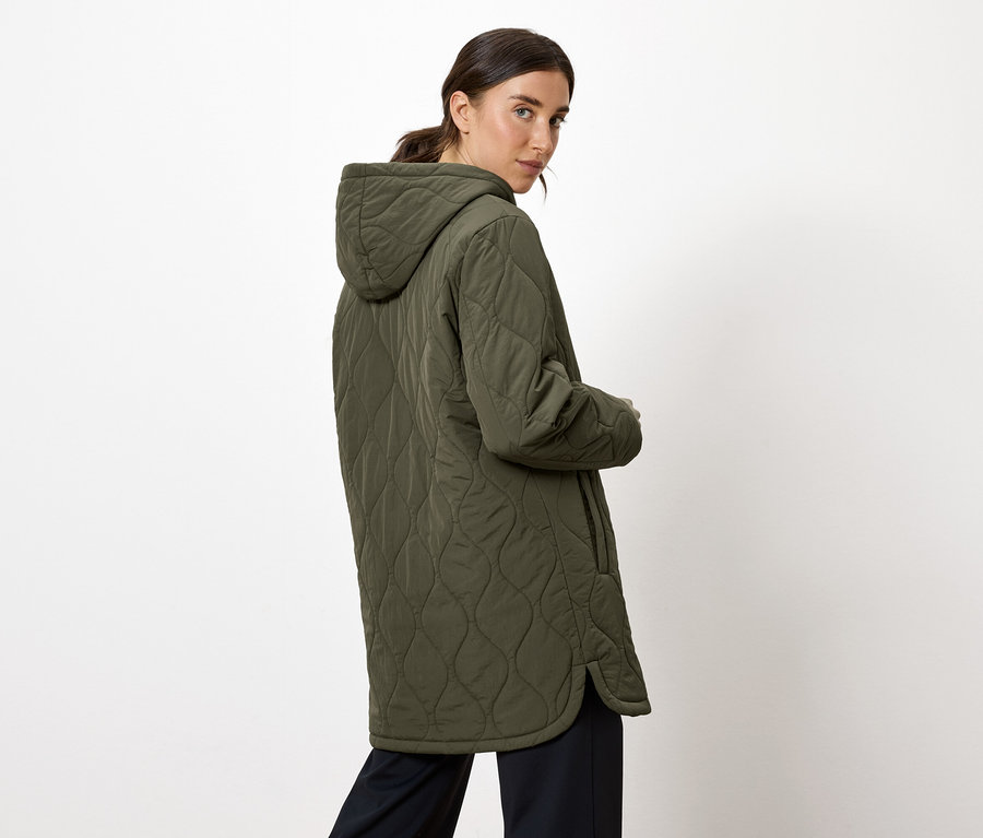 Žena sa pozerá cez plece a má na sebe olivovozelený prešívaný kabát s kapucňou.