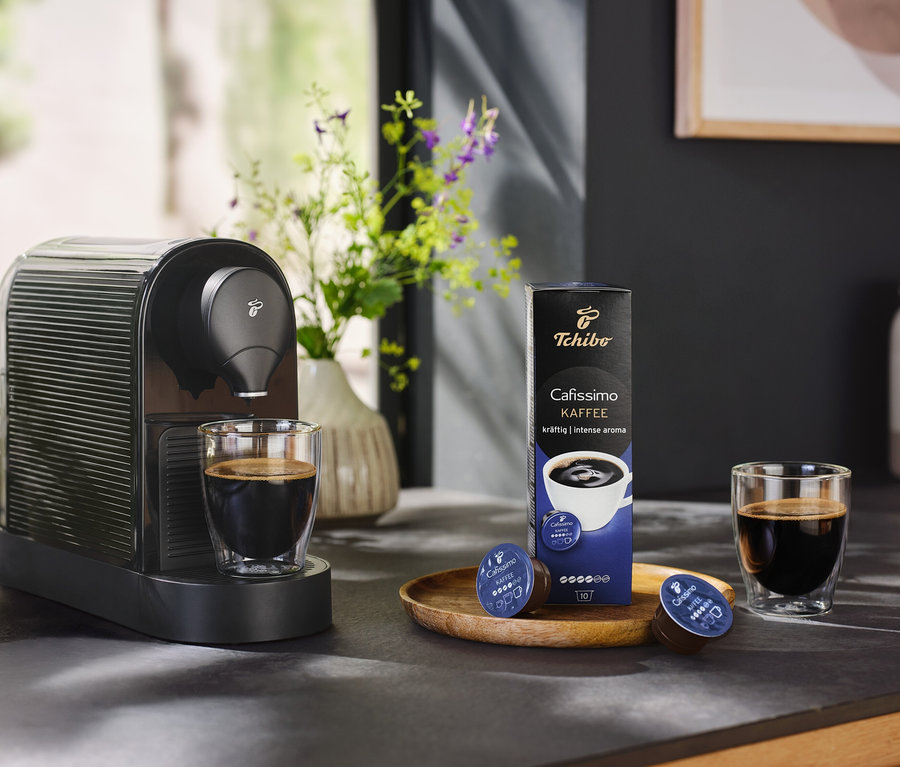 Čierny kávovar Cafissimo pure+ s šálkou kávy a balením kávových kapsúl.