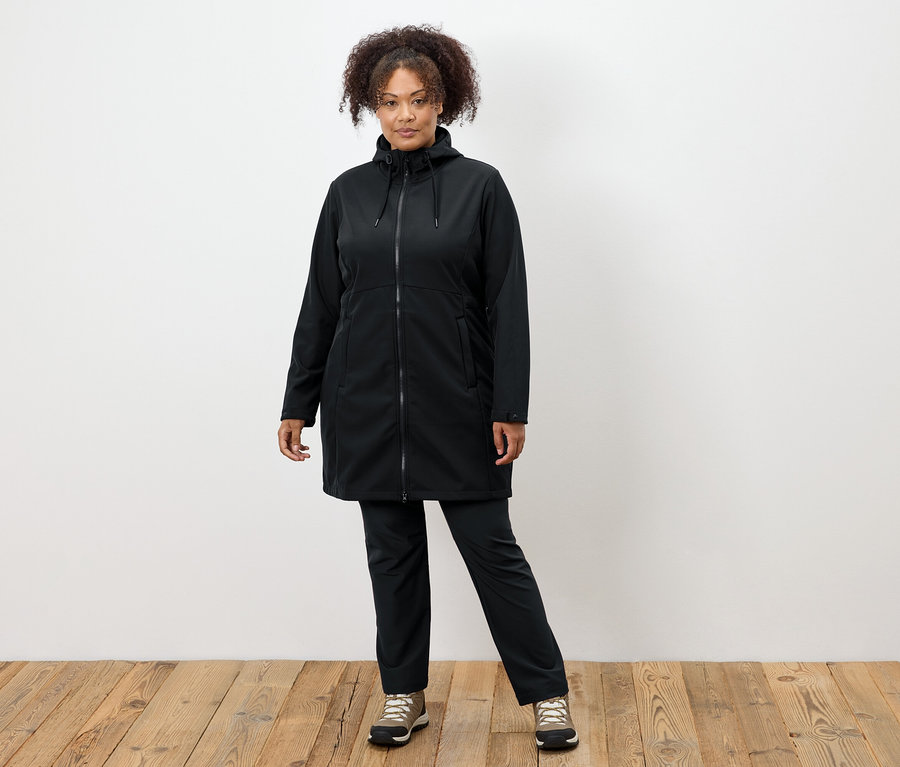 Žena v čiernom kabáte a nohaviciach stojí na drevenej podlahe.