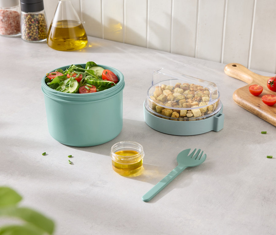 Miešaný šalát v tyrkysovej nádobe s krutónmi v nádobe s priehľadným vekom a dresingom a vidličkou vedľa.