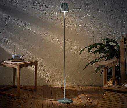 Šalviová LED akumulátorová stojaca lampa do exteriéru stojí vedľa dreveného stolíka so šálkou a dreveného kresla.