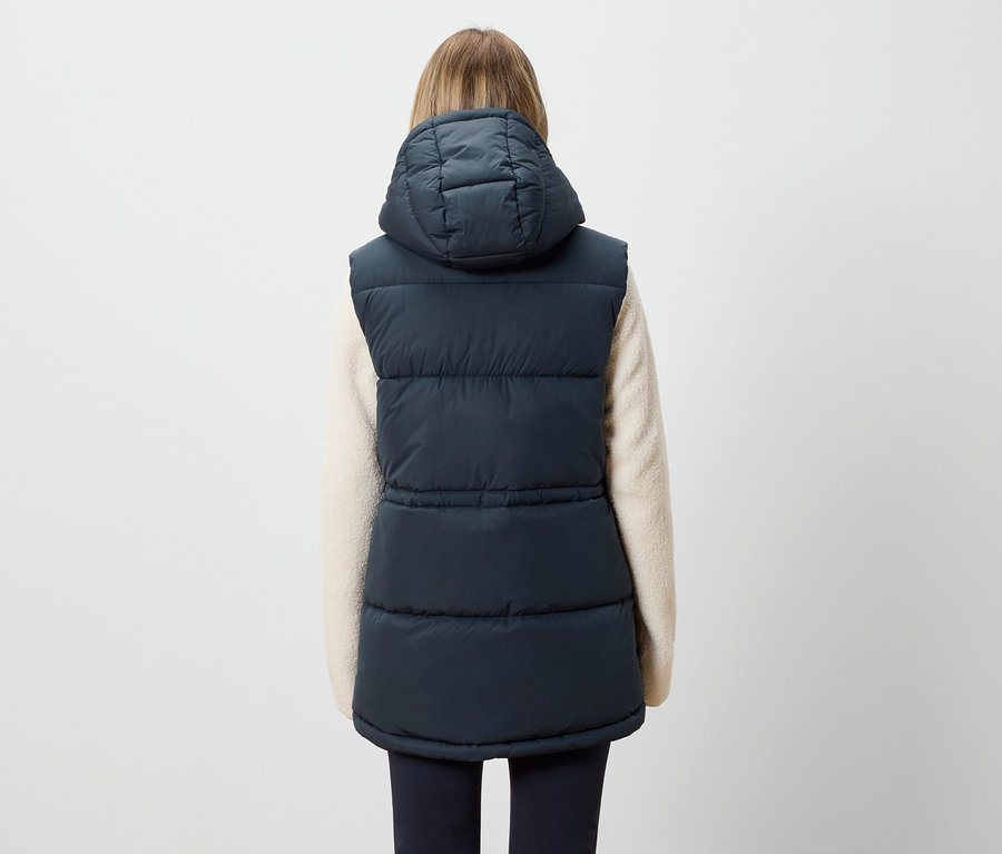 Zozadu je vidieť žena s blond vlasmi, ktorá má na sebe tmavomodrú prešívanú vestu s kapucňou a krémový sveter.