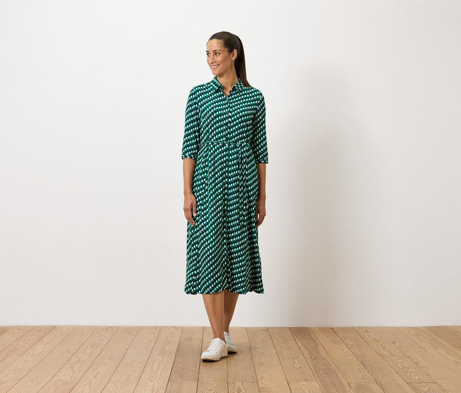 Žena stojí a má na sebe zelené vzorované šaty a biele tenisky.