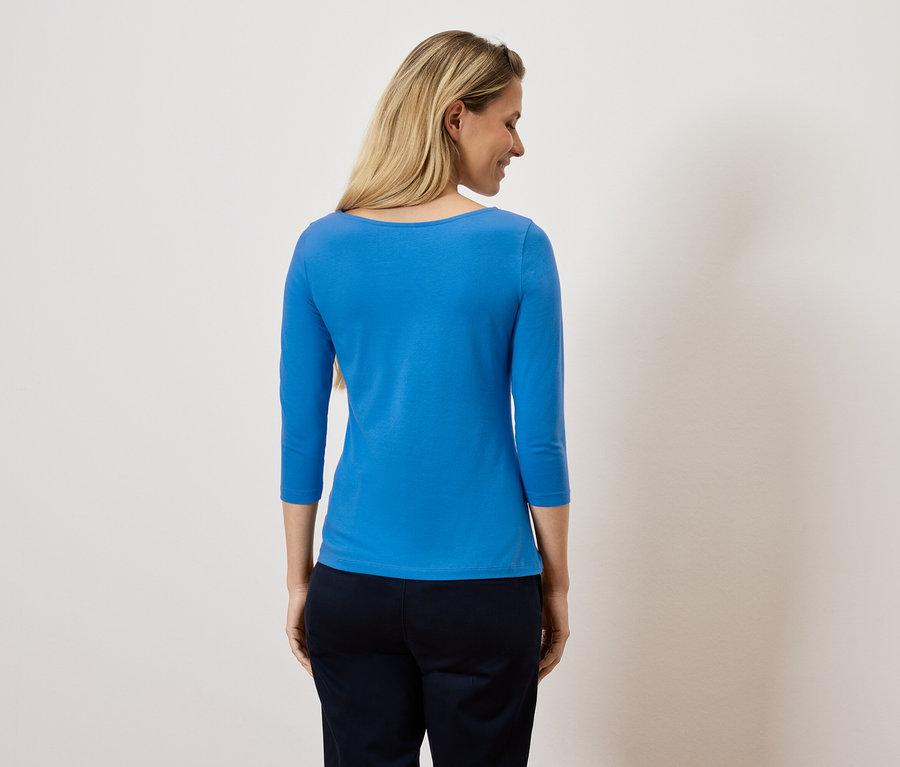 Žena sa pozerá cez rameno a má na sebe modrý top s trojštvrťovým rukávom a tmavé nohavice.