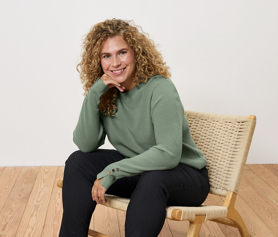 Žena sedí na stoličke v zelenom svetri a čiernych nohaviciach.