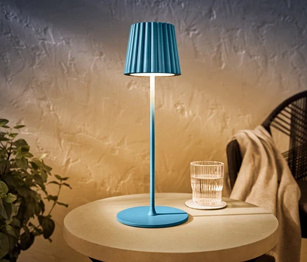 Modrá LED akumulátorová stolová lampa do exteriéru stojí na stole vedľa pohára vody.