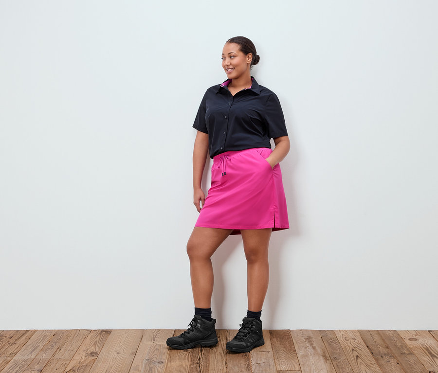 Žena stojí pred bielou stenou, má na sebe čiernu košeľu, ružovú sukňu a čierne topánky.