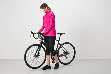 Žena v ružovej bunde, čiernych cyklistických nohaviciach a čiernych topánkach stojí vedľa čierneho bicykla.
