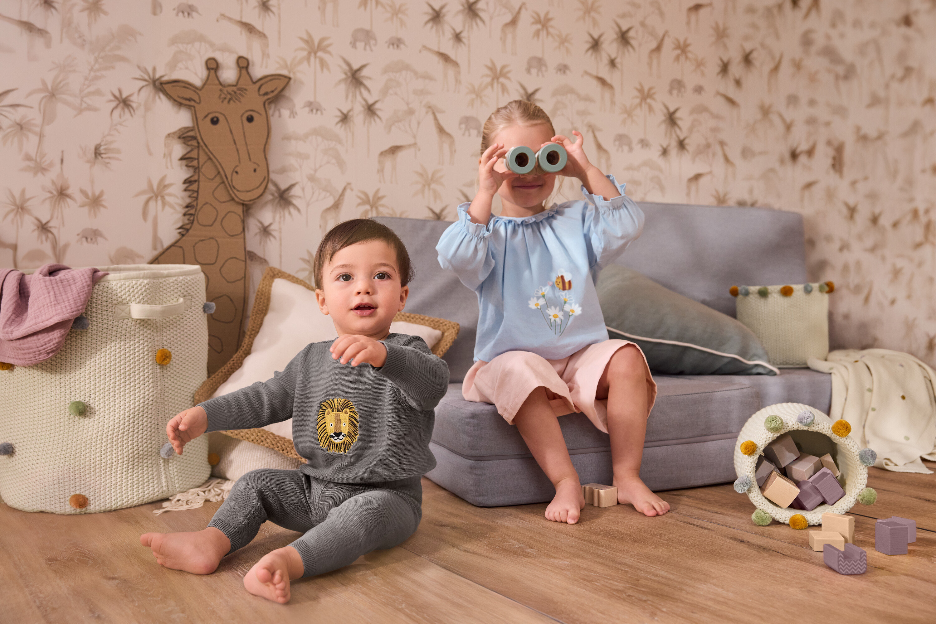 Batoľa sedí vedľa veľkého úložného koša do detskej izby. Dievča sedí na sivej pohovke a drží si pred tvárou pletenú deku pre bábätká. Má na sebe detskú šambré blúzku a detské menčestrové culottes nohavice.