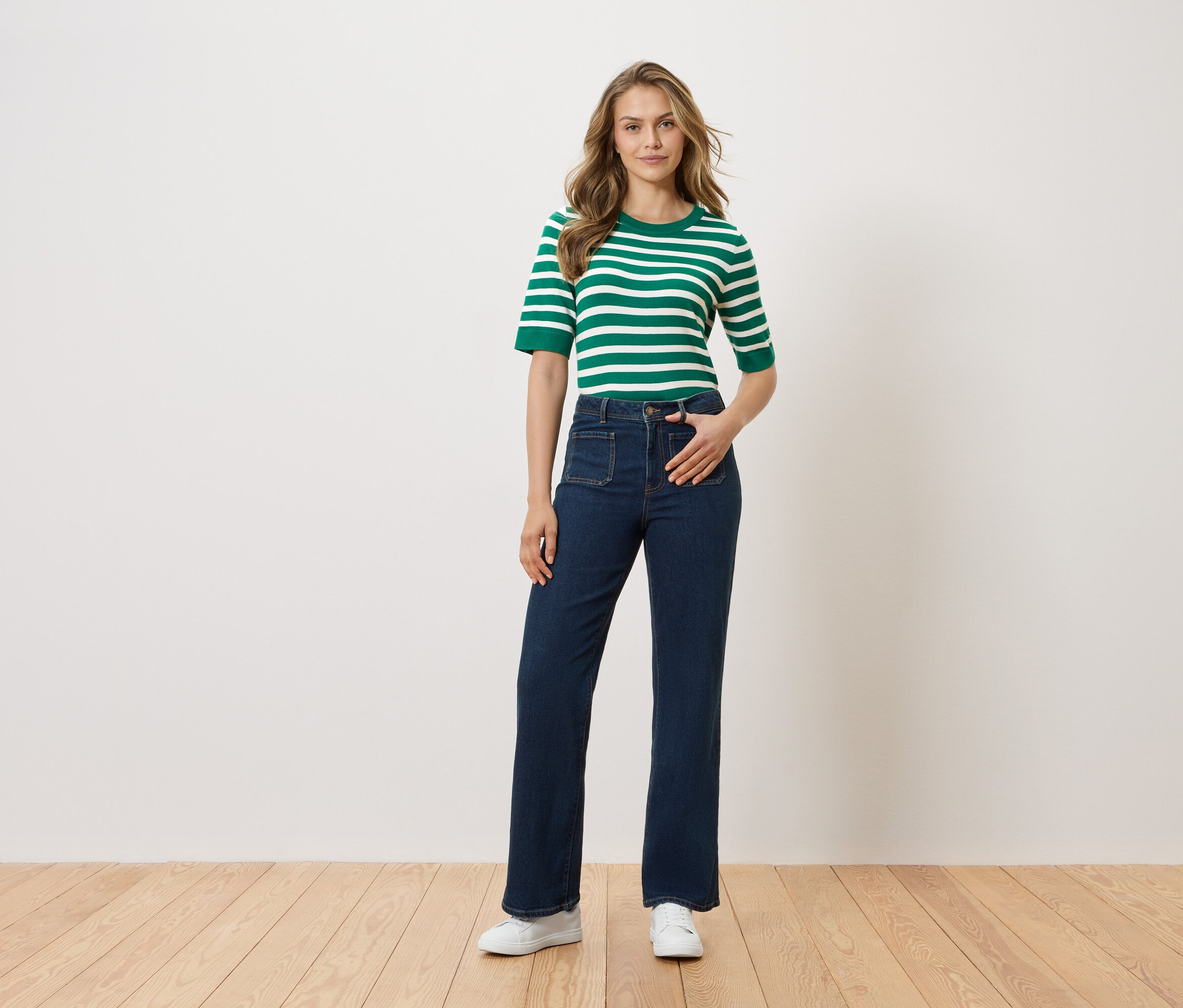 Žena stojí s rukou vo vrecku nohavíc pred bielou stenou. Má na sebe zeleno-biely pruhovaný top, tmavé džínsy a biele topánky.