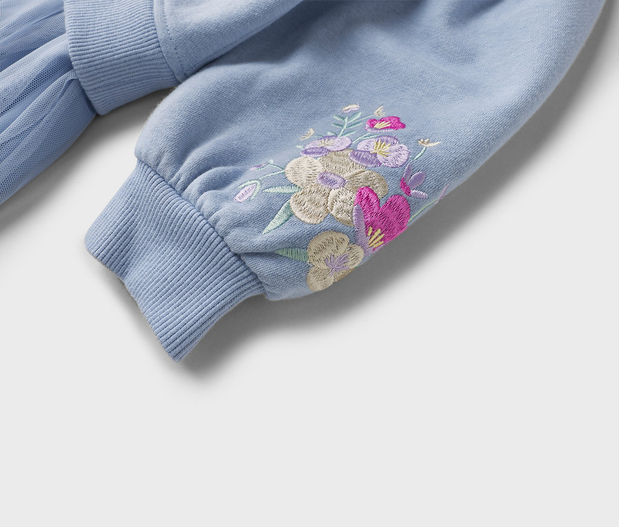 Detail svetlomodrej mikiny s kvetinovou výšivkou na rukáve a rebrovaným lemom.