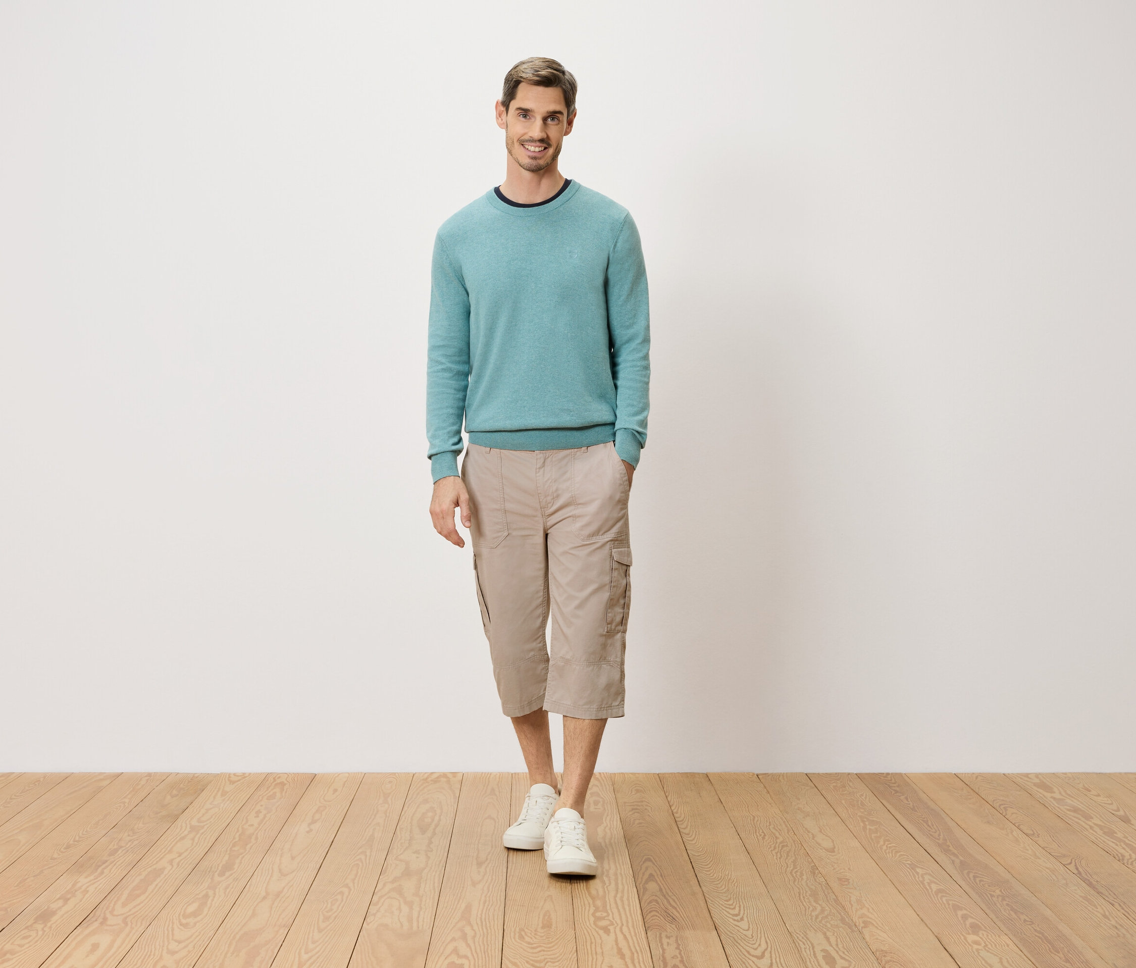 Muž stojí s rukou vo vrecku pred bielou stenou. Má na sebe svetlomodrý sveter, béžové trojštvrťové cargo nohavice a biele topánky.