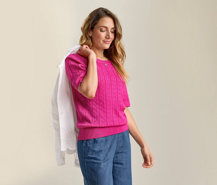 Žena s Jeansjacke mit Lochstickerei prehodenou cez rameno a pulóvrom s vrkočovým vzorom z jemnej pleteniny v ružovej farbe a s džínsami.