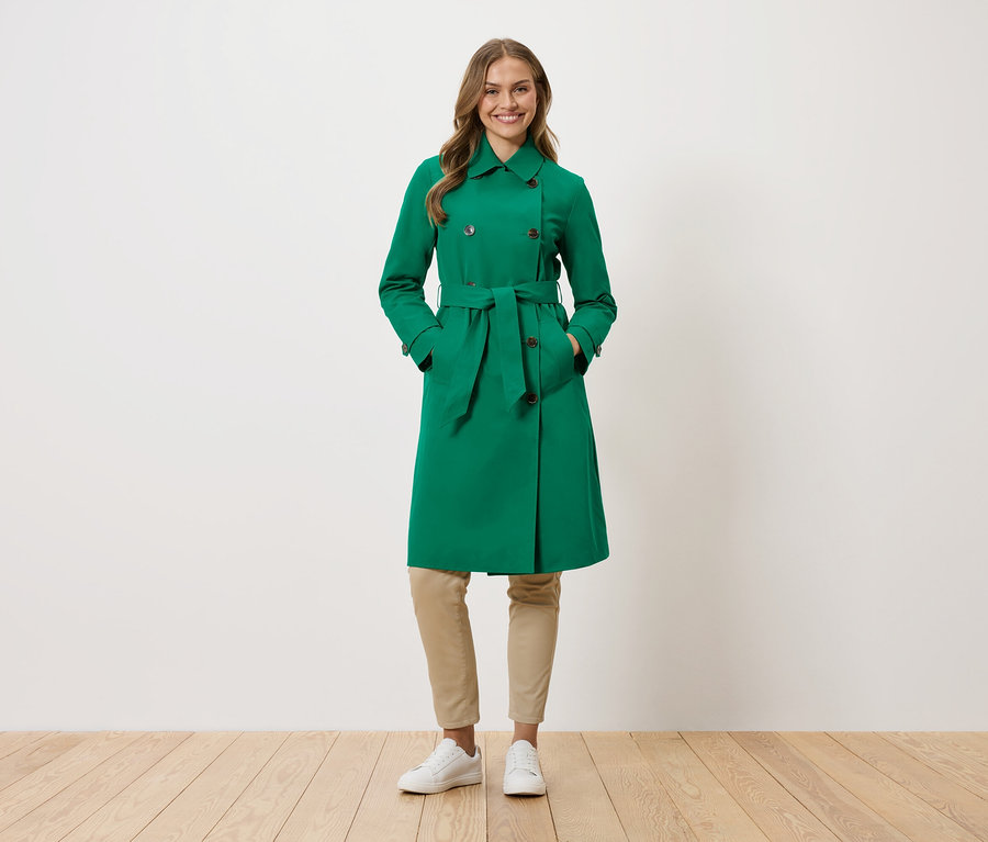 Žena stojí s rukami vo vreckách zeleného trenčkotu.