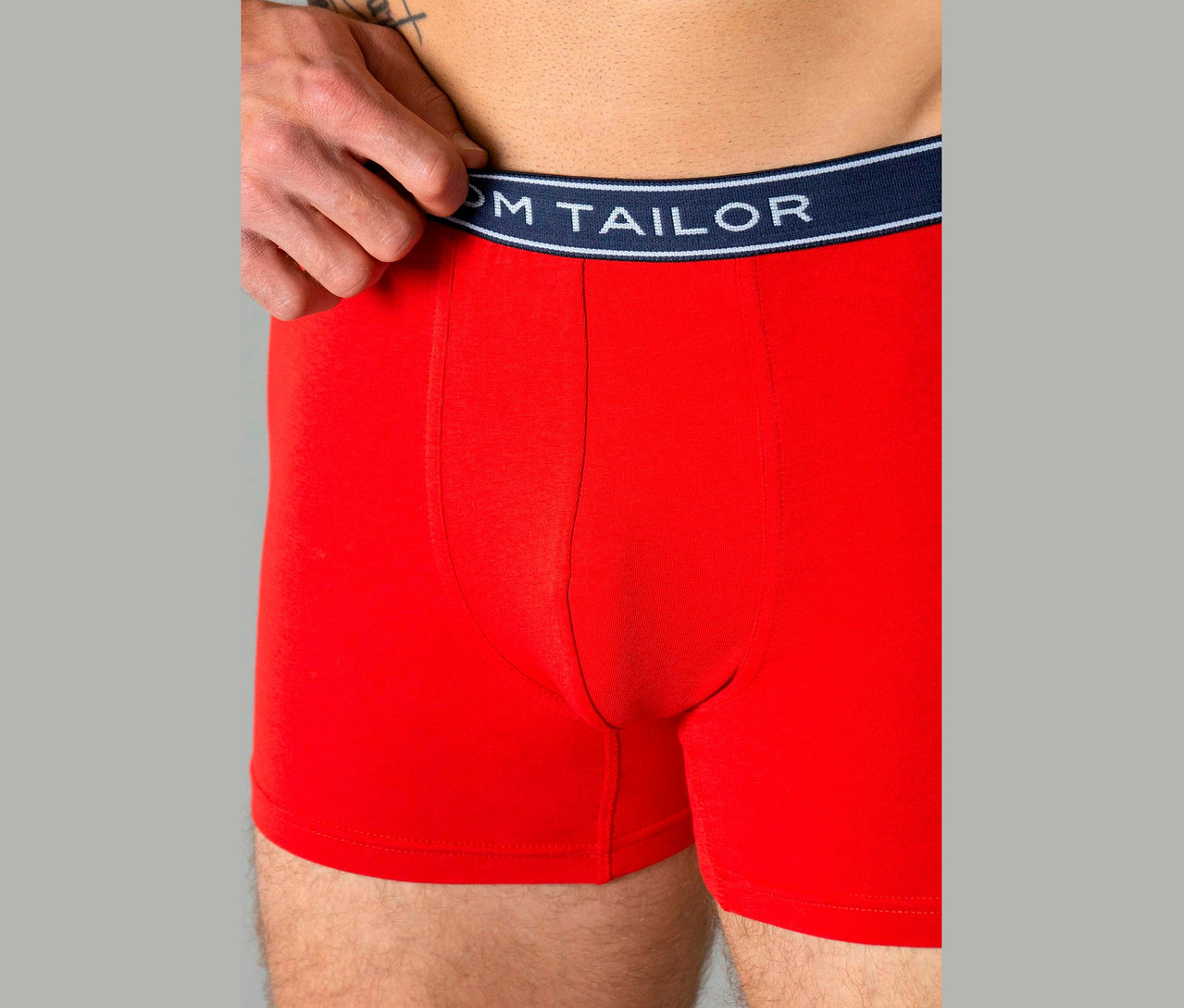 Muž má na sebe červené boxerky s tmavomodrým pásom s nápisom „TOM TAILOR“.