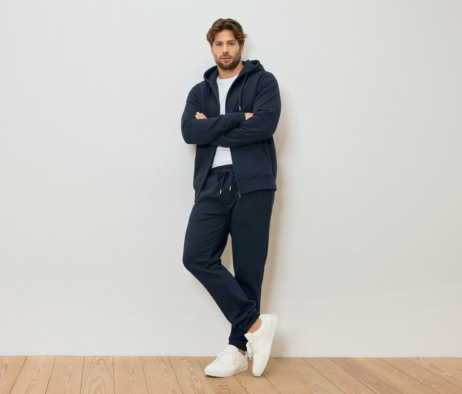 Muž so založenými rukami má na sebe tmavomodrú mikinu s kapucňou na zips, tmavomodré tepláky a biele tenisky, stojí pred bielou stenou.