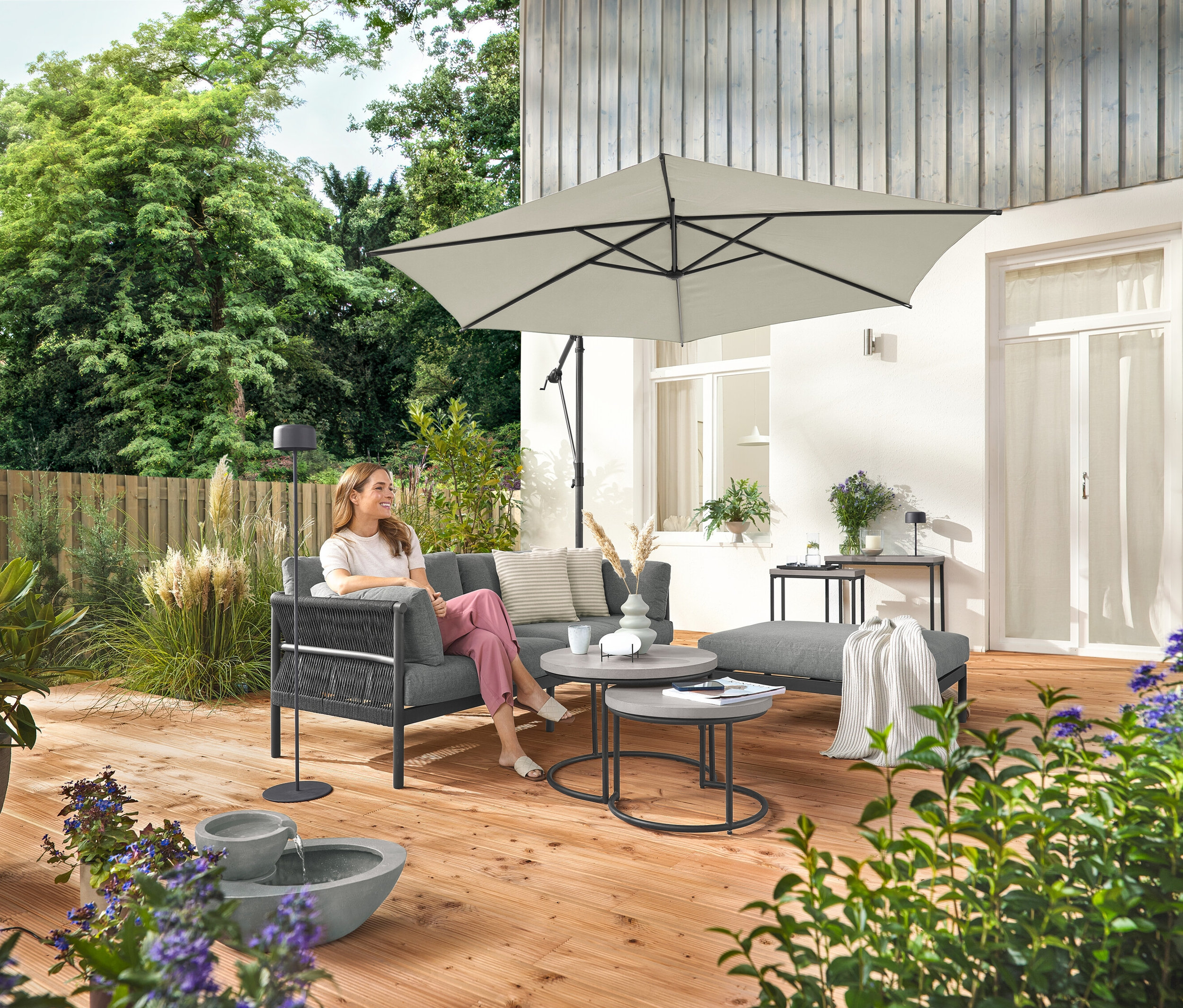 Žena sedí na komfortnej záhradnej pohovke so samostatnou podnožkou. Vedľa nej stojí sivá solárna stojacia lampa. V popredí je solárna záhradná fontána.