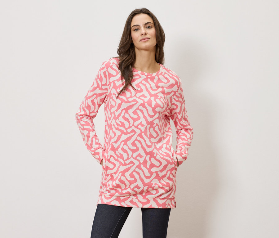 Žena s rukami vo vreckách má na sebe ružový vzorovaný top s dlhým rukávom a tmavé džínsy.