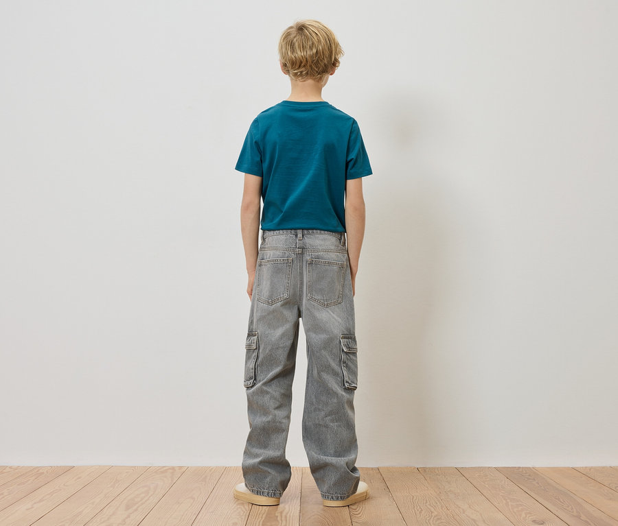 Chlapec s blond vlasmi stojí chrbtom ku kamere, má na sebe modrozelené tričko a sivé cargo džínsy.