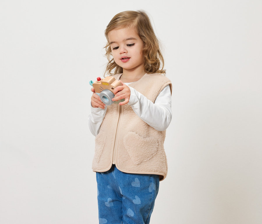 Malé dievča s kučeravými vlasmi drží hračkársky fotoaparát na svetlom pozadí.